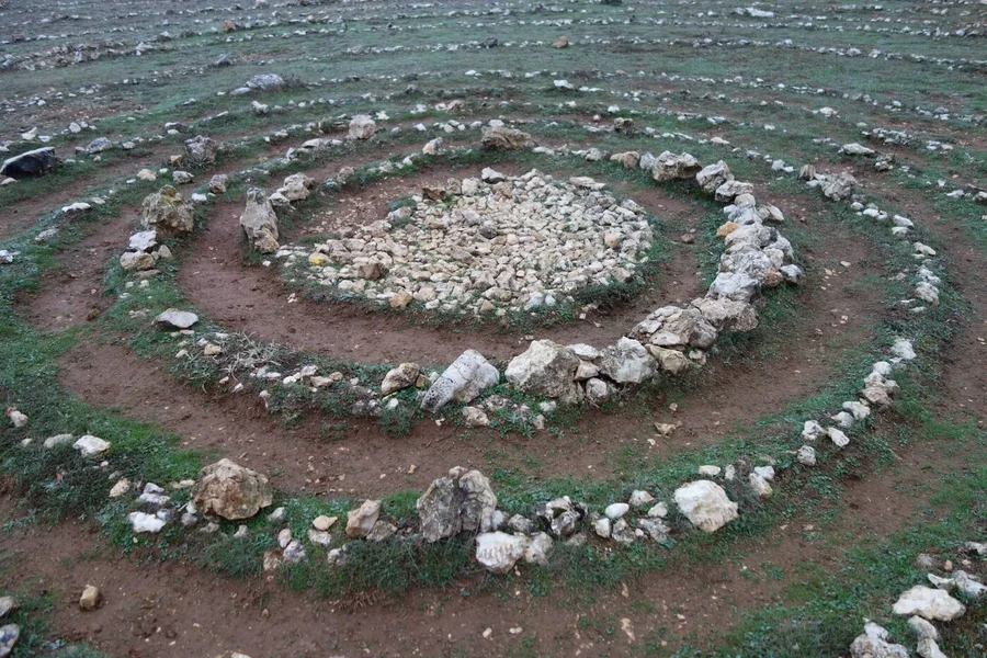 Eine große Spirale aus hellen Steinen ist in eine karge Wiese gelegt. In der Mitte liegt ein Kreis aus kleineren Steinen.