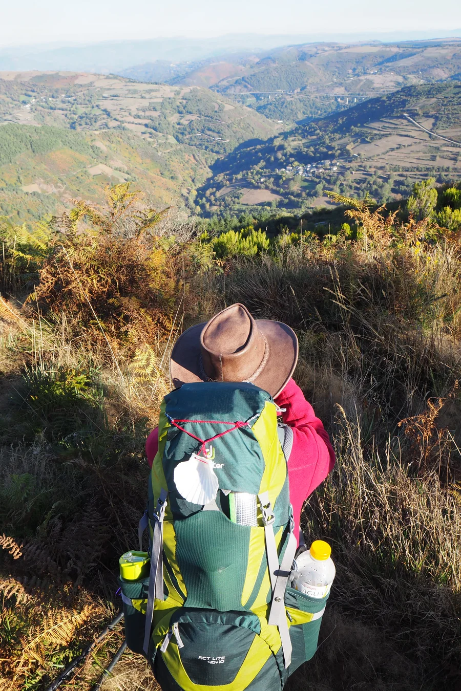 Pilgernde Person mit Rucksack blickt von einer Höhe in ein weites Tal