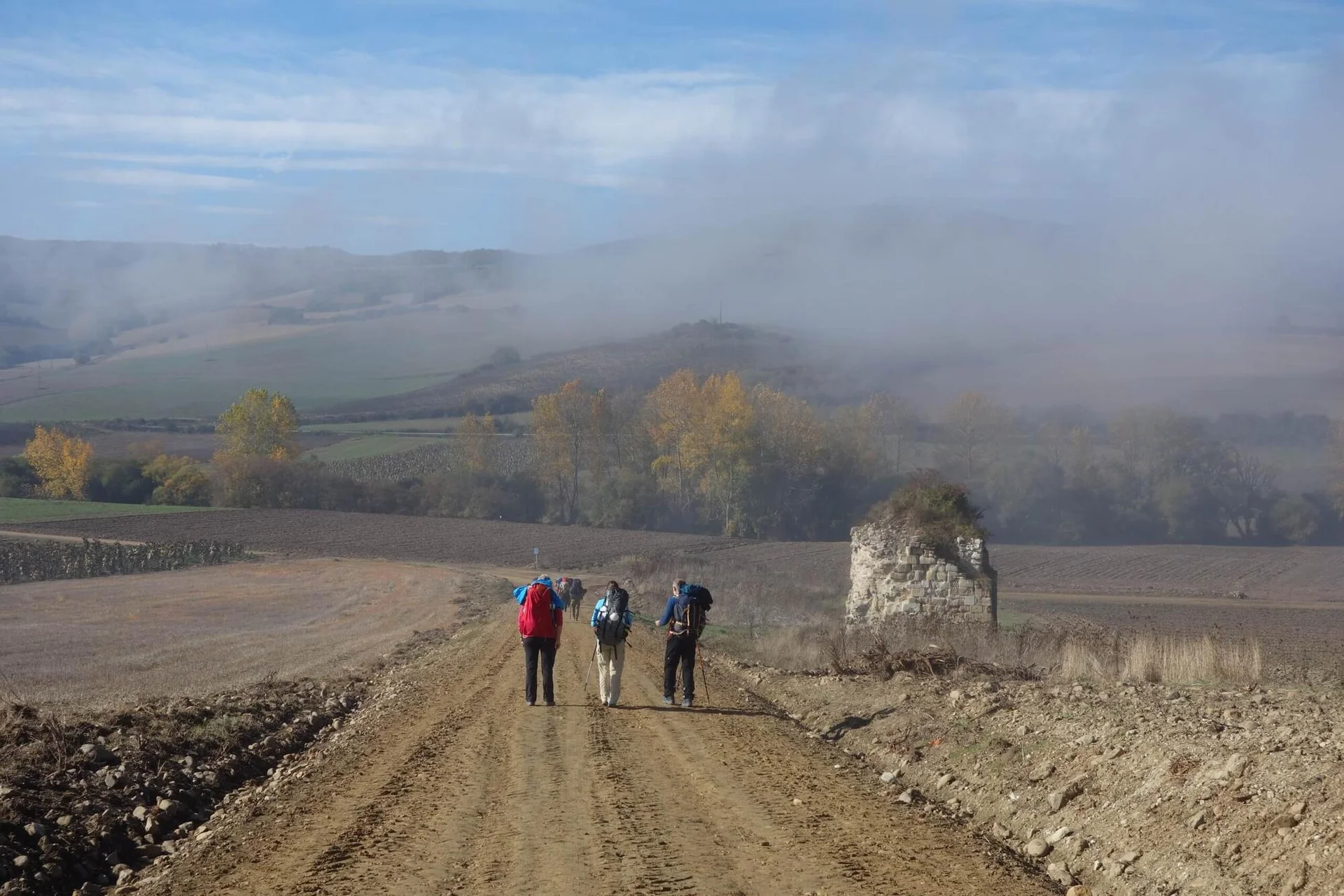 Ein breiter Feldweg führt durch herbstliche Felder auf eine nebelverhangene Hügelkette zu. In der Ferne gehen drei Menschen mit Rucksäcken.
